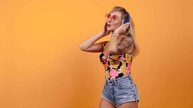 Sexy hot woman listening and enjoying music in headphones starts dancing isolated on yellow orange background in studio. She is wearing sunglasses. Cheerful, pretty and attractive girl enjoys life