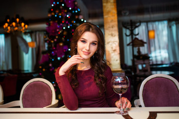 Beautiful young woman in gray dress stands with wine glass in her hands on background of Christmas tree, lights and garlands.