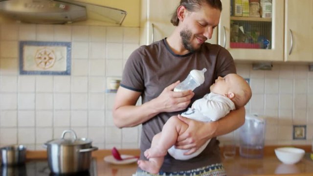 A young multitasking father and an infant in the kitchen. The man finishes feeding the baby, takes the bottle and both smile. Man and child in a modern kitchen