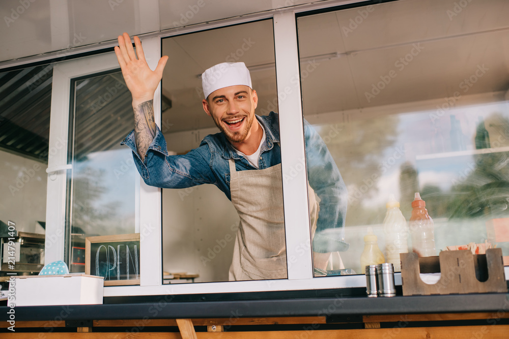 Sticker cheerful young man waving hand and smiling at camera while working in food truck