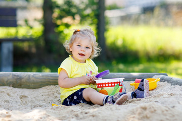 Cute toddler girl playing in sand on outdoor playground. Beautiful baby in red gum trousers having fun on sunny warm summer day.