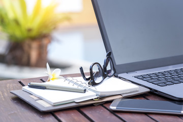 Laptop with smart phone empty notebook pen and glasses on wooden table near swimming pool.