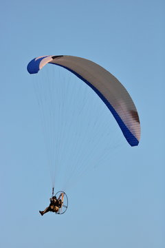 Paralotniarz lata w powietrzu, w posyzcji siedzącej, widać śmigła silnika, czasza lotni biało-niebieska, błękitne czyste niebo