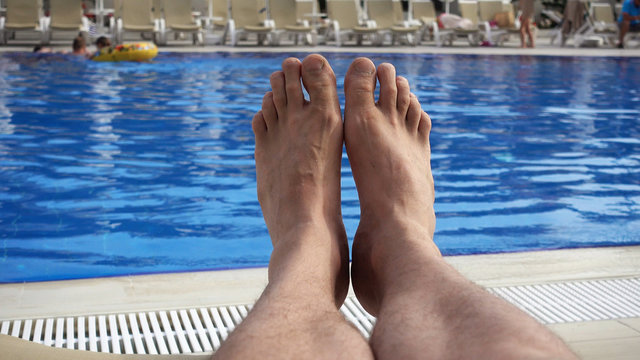 A first person view, a point of looking at a man who is resting by the pool. The legs of a man who is sunbathing near the pool and looks ahead of himself.