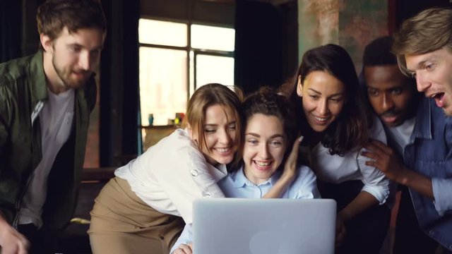 Young woman is looking at laptop screen and laughing, her colleagues are coming to her, hugging and congratulating her doing high five. Success and business concept.