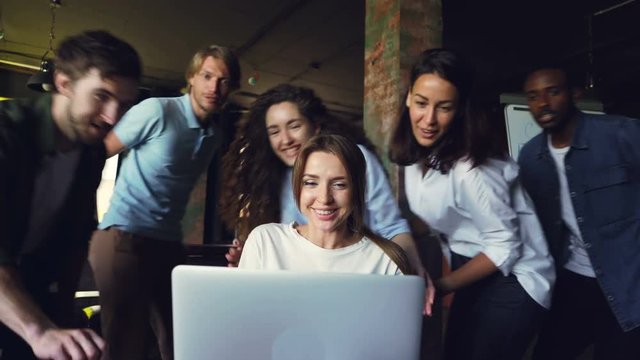 Attractive young office worker is happy about good work results, she is looking at laptop screen and laughing, her coworkers are clapping hands and hugging her.