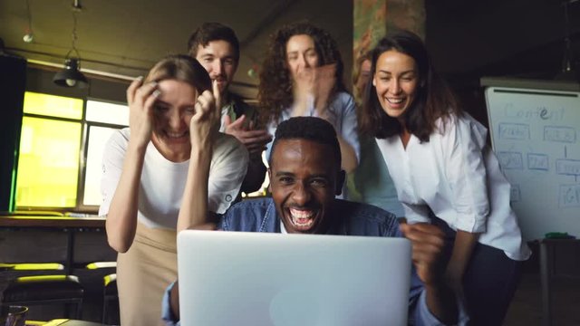 African American man office worker is excited about good news, he is looking at laptop screen and laughing, his team is celebrating success, clapping hands and congratulating him.