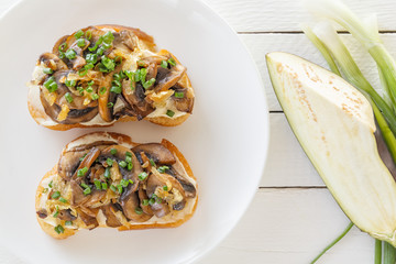 Two fresh bruschettes with mushrooms on lie on a plate next to a peeled eggplant and onions on a white wooden background. Top view