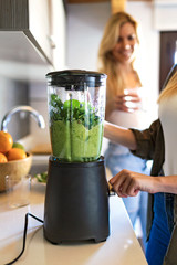 Beautiful young woman preparing detox juice in the blender and talking with friend at home.