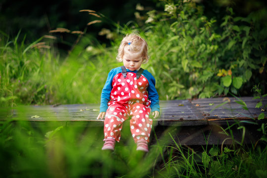 Cute adorable toddler girl sitting on wooden bridge and throwing small stones into a creek. Funny baby having fun with outdoor games in nature.