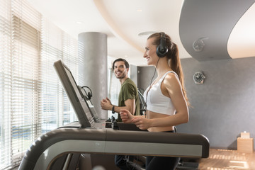 Happy young woman listening to music while running beside a handsome man on a modern treadmill with...