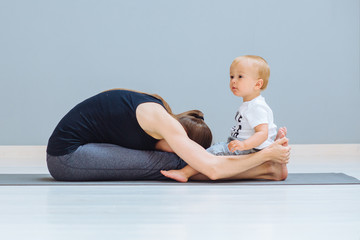 Sportive brunette mother with baby son relax after good workout on grey yoga mat over gray wall...