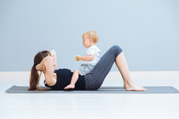 Obraz premium A sports mother is engaged in fitness and yoga or pilates exercie with a toddler son at home. Child sitting on mom's abdomen. Empty gray background. Motherhood, simple healthy lifestyle concept.