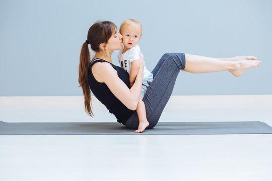 Mom swings a press and kissing baby. A sports mother is engaged in fitness pilates exercie with a toddler son at home. Child sit on mom's abdomen and looks at you. Motherhood, healthy lifes concept.