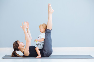Sportive brunette mother with baby son doing press exercise on grey yoga mat over gray wall background. Athletic and healthy motherhood. Fitness, happy maternity and healthy lifestyle concept.
