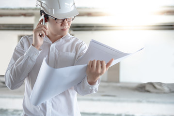 Asian male engineer or architect holding blueprints or architectural drawing while using smartphone for calling at construction site. Engineering, Architecture and building construction concepts
