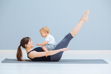 Sportive brunette mother with baby son doing press exercise on grey yoga mat over gray wall background. Athletic and healthy motherhood. Fitness, happy maternity and healthy lifestyle concept.