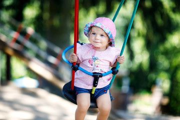 Cute adorable toddler girl swinging on outdoor playground. Happy smiling baby child sitting in chain swing. Active baby on sunny summer day outside