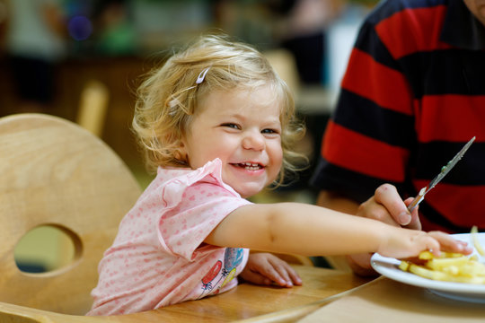 Adorable Toddler Girl Eating Healthy Vegetables And Unhealthy French Fries Potatoes. Cute Happy Baby Child Taking Food From Parents Dish In Restaurant