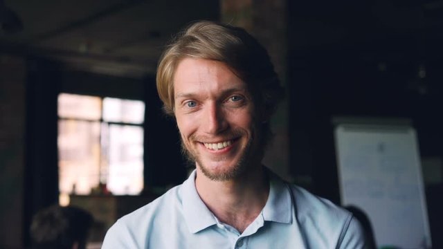 Close-up slow motion portrait of happy young man entrepreneur standing in workplace, looking at camera and smiling. Modern office with people is in background.