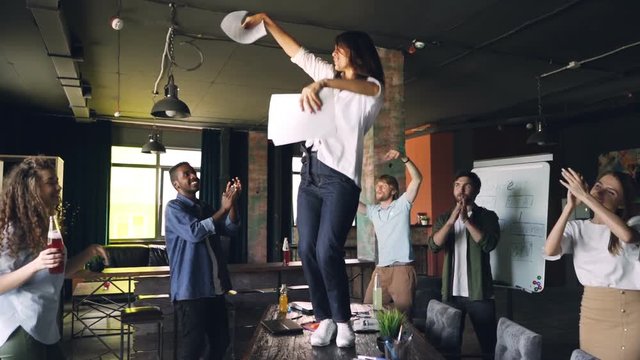 Clow motion of joyful young lady dancing on table at office party with pile of paper and throwing documents while her team is laughing and dancing around her.
