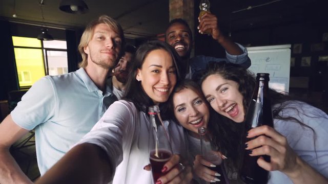 Slow motion portrait of group of people coworkers taking selfie with drinks at office party, attractive men and women are posing with funny faces, holding bottles and laughing.