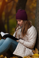 Young female student wearing coat and knitted hat sitting in the fall park with book in hands