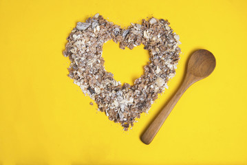 Heap of oat flakes in a shape of heart isolated on yellow background with wooden spoon.