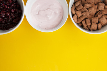 Bowls of whole grain muesli, dried fruits, granola oat meals and yogurt cream or milk, arranged in line, with copy space. Isolated on yellow background. Top view.