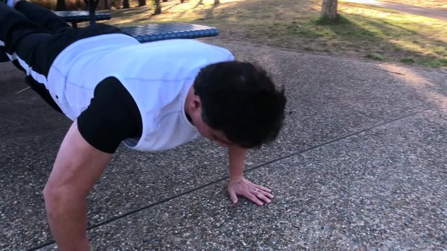 A man,runner, does push ups by the park bench.