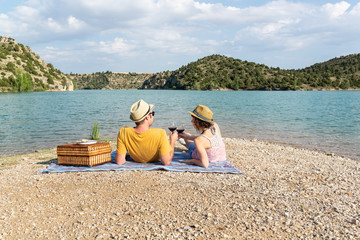 Pareja joven brindando y mirándose el uno al otro durante un romántico picnic cerca de un hermoso lago