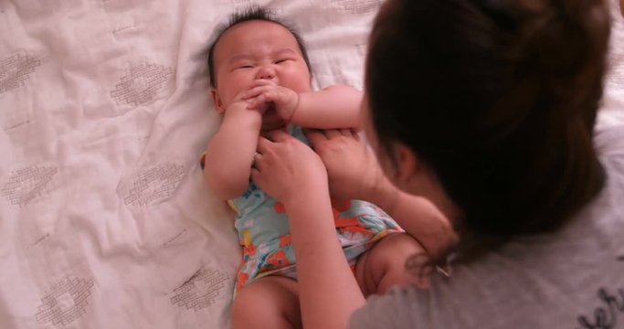 Asian mother is tickling her baby girl on the bed, baby is eating her fingers