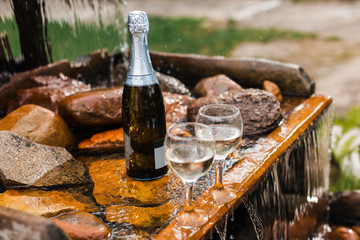 Love, romance, holiday, celebration concept. Bottle and glasses of champagne chilled by waterfall in summer forest on sunset.