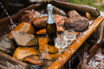 Love, romance, holiday, celebration concept. Bottle and glasses of champagne chilled by waterfall in summer forest on sunset.