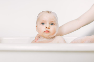 Happy laughing baby taking a bath playing with foam bubbles. Little child in a bathtub. Infant washing and bathing.