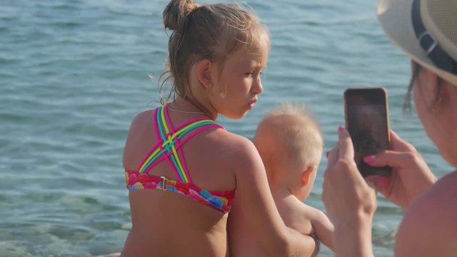 Happy mother taking picture of her children with smartphone on the beach outdoors. Mother taking picture of her family at the sea. Vacation concept.