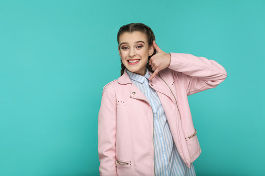 call me gesture. portrait of beautiful cute girl standing with makeup and brown pigtail hairstyle in striped light blue shirt pink jacket. indoor, studio shot isolated on blue or green background.