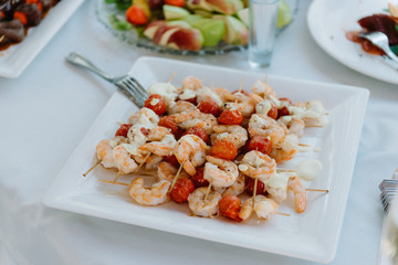 The snack made on a grill. Grilled shrimp on skewers on a white board with lime and tomatoes