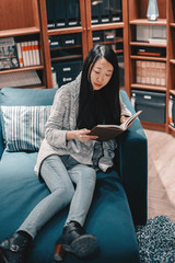 Full length portrait of beautiful young asian girl in jeans and coat sitting in blue armchair reading a book
