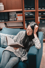 Obraz premium Half length portrait of beautiful young asian girl in jeans and coat sitting in blue armchair reading a book
