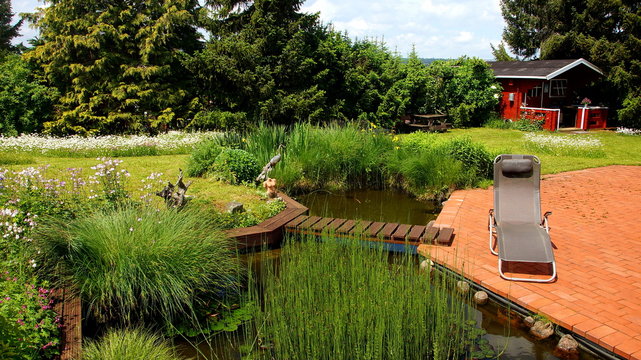 attraktiver naturbelassener Teich mit Brücke an rote Terrasse angrenzend und Gartenhaus