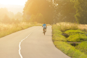 A young guy rides a bicycle on an asphalt road, leaves in the distance, the evening sunset