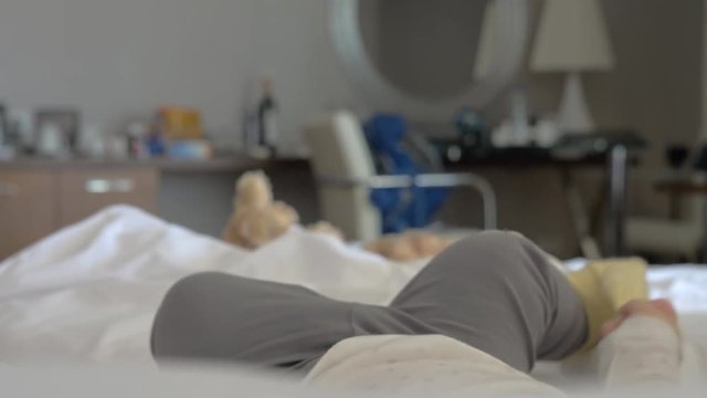Little baby girl (with no visible head) is lying on her back on a bed and playing with her feet. She is wearing grey comfy pants with pink hearts on the knees and yellow socks one of which she is