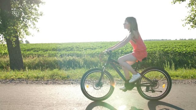 Side full length view of beautiful teenage girl biking through fields during sunset in summer. Schoolgirl enjoying summer holidays while riding bike in countryside. Sun flare through trees. Steadicam