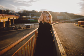 Beautiful young hipster woman with blonde hair in black fur coat outdoors on the road on sunset. Grunge. Urban street style.