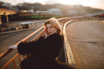 Attractive blonde hipster woman with colorful make-up in black fur coat posing outdoors on the road on sunset. Urban background. Grunge style