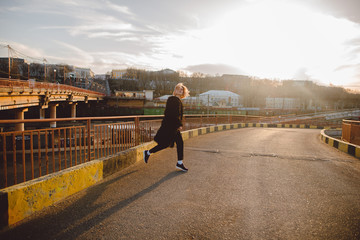 Fototapeta premium Attractive blonde hipster woman in black fur coat running outdoors on the road on sunset. Urban background. Grunge street style