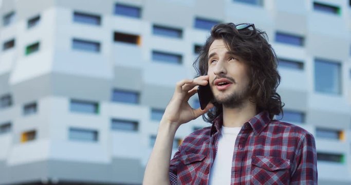 Close up of the young attractive man with long hair and a plaid shirt talking on the phone at the big modern building background. Outdoor.
