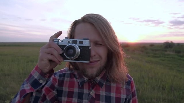 Photographer taking photo with retro camera. Hipster with long curly hair and plaid shirt taking pictures in outdoor on sunset and smiling. Slowmotion.