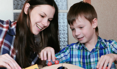 Playing together. Happy Mom and son are playing a wooden railway with train, wagons and tunnel sitting at the table.